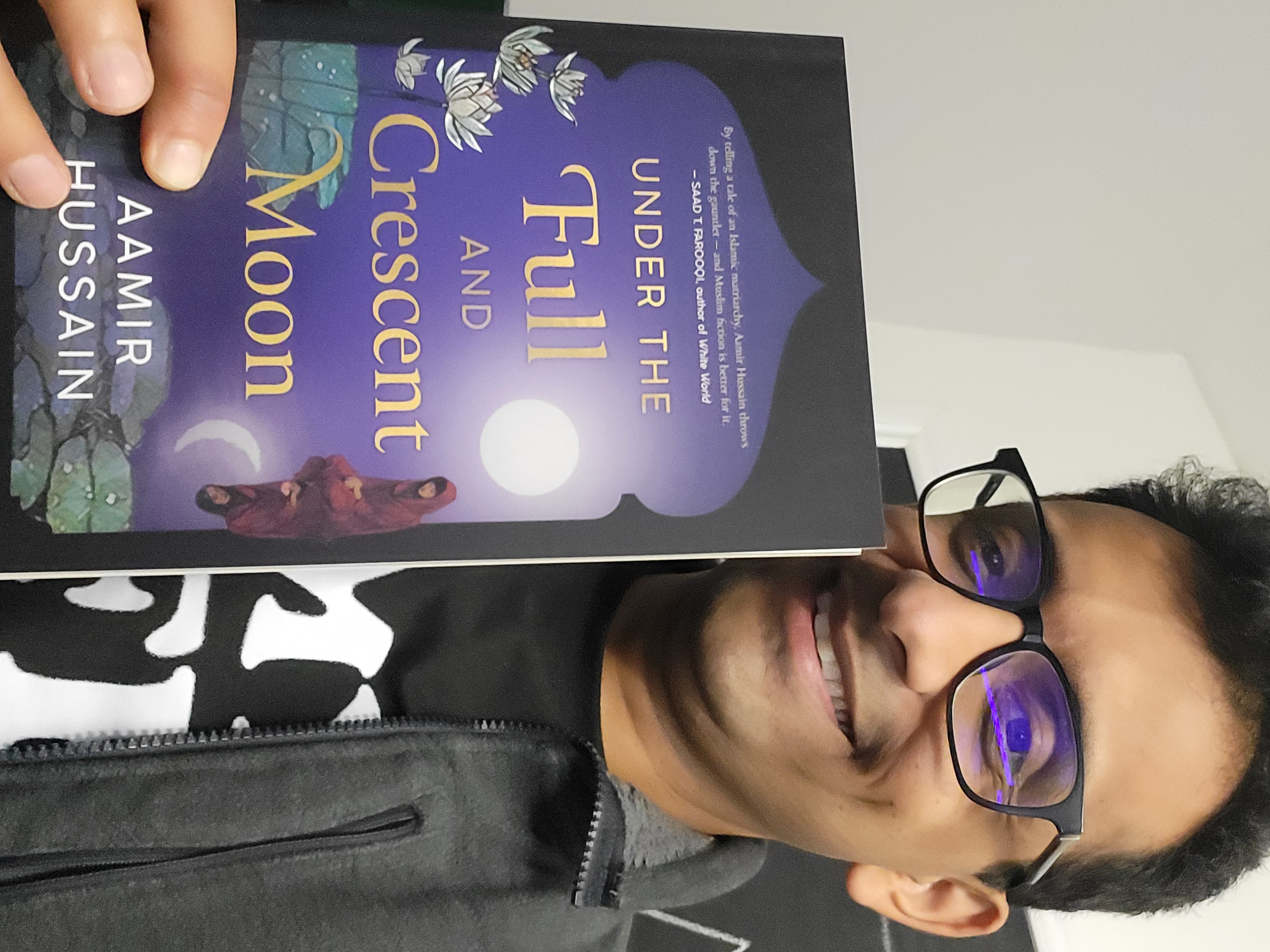 Picture of Aamir Hussain, the author of "Under the Full And Crescent Moon" Holding up a print copy of the book which shows a young woman wearing a hijab kneeling in front of a pool of water, above her is a full moon, reflected in the water is her without a hijab and a crescent moon 