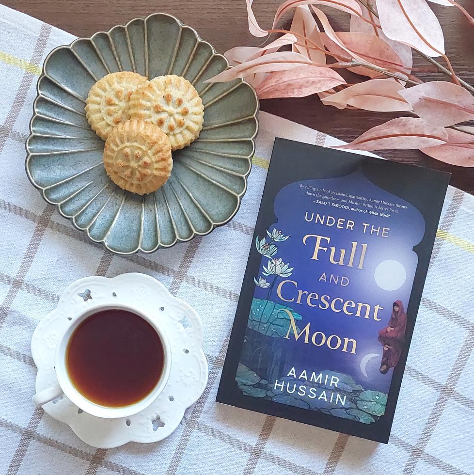 Artistic Picture of the novel "Under The Full And Crescent Moon" next to a cup of tea or coffee and a plate of Ma'amoul date pastries on top of a checkerboard tablecloth and next to pink leaves on a brown table